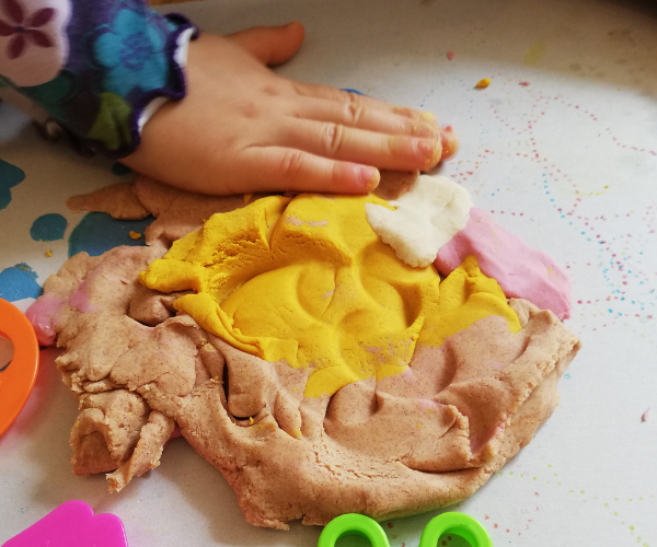 Kinderhand, die knetet und Knete mit Förmchen aussticht. Die Knete ist gelb und rosa gefärbt.